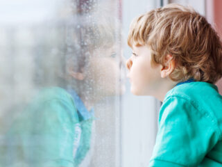 Lovely little blond child sitting near window and looking at raindrops, indoors.